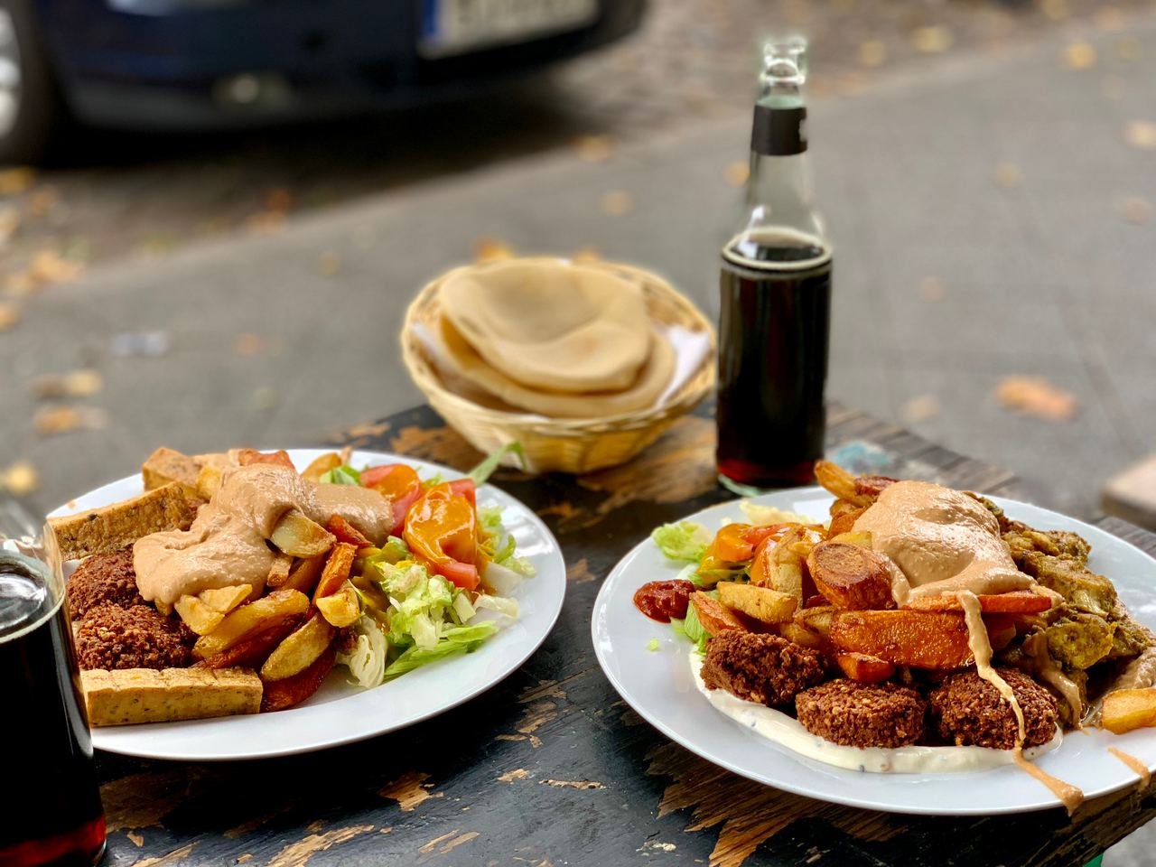 Zwei weiße Teller, jeweils mit Falafel, Halloumi und Salat. Im Hintergrund rundes arabisches Brot und eine Flasche Fritzcola.