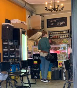 Innenraum des Dar4 Bistros. Zu sehen sind schwarze Stühle an beiden Seiten, Getränkekisten in der Mitte vor einer Theke. Davor steht eine Frau beim bestellen, Jeans und grünes Langarmshirt.