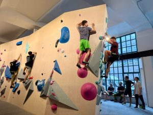 "Das Elektra" Boulderhalle - Wände mit bunten Bouldern, Menschen bouldern an der Wand und stehen am Rand und sehen zu.