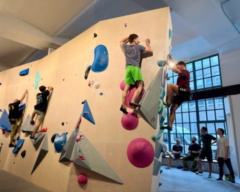 "Das Elektra" Boulderhalle - Wände mit bunten Bouldern, Menschen bouldern an der Wand und stehen am Rand und sehen zu.