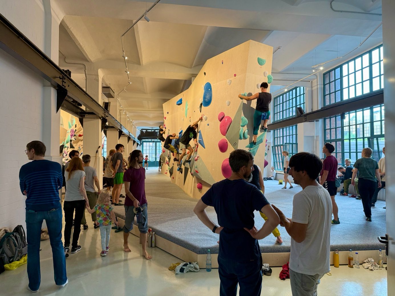 "Das Elektra" Boulderhalle - Wände mit bunten Bouldern, Menschen bouldern an der Wand und gehen durch die Halle